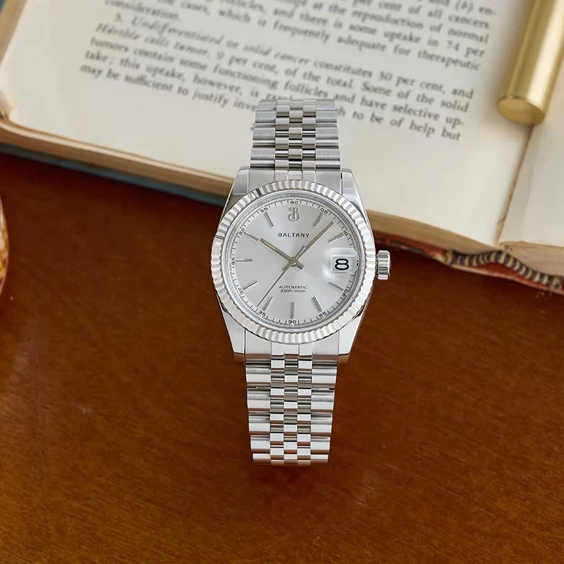 NorthChrono silver automatic watch with a round black dial, date display, textured bezel, and metal bracelet, displayed on a wooden surface beside an open book and a golden object.