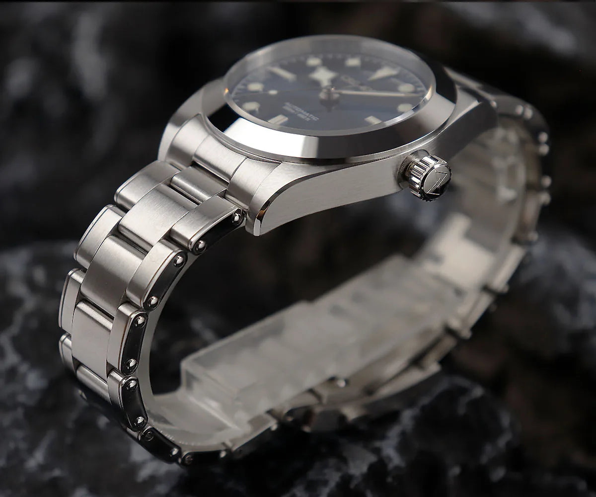 NorthChrono close-up of a sporty wristwatch featuring a black dial with metallic hour markers, a round stainless steel case, and a stainless steel bracelet. A crown is visible on the side against a textured stone background.