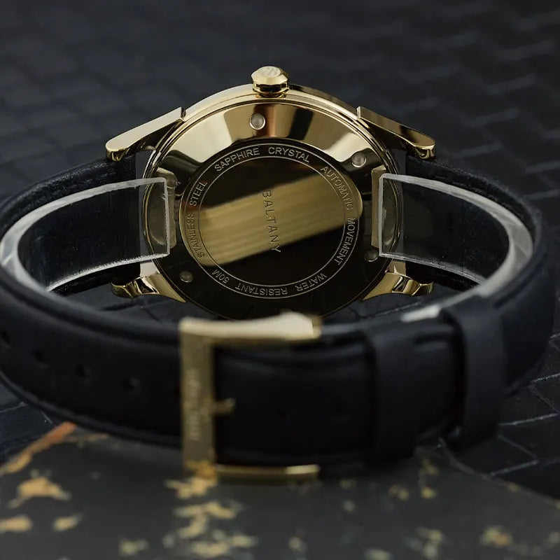 NorthChrono timepiece showcasing the back view of a gold-toned case with markings for "sapphire crystal" and "water resistant." Features a black strap on a textured leather background, highlighting its sleek design.