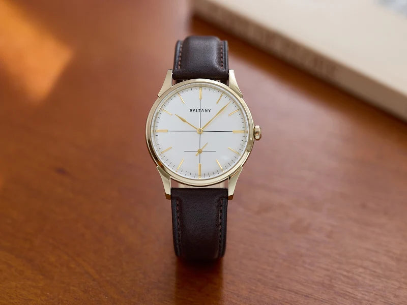 NorthChrono automatic watch with a white dial featuring gold-toned hour markers and hands, paired with a brown leather strap, placed on a wooden surface with a book partially visible in the background.