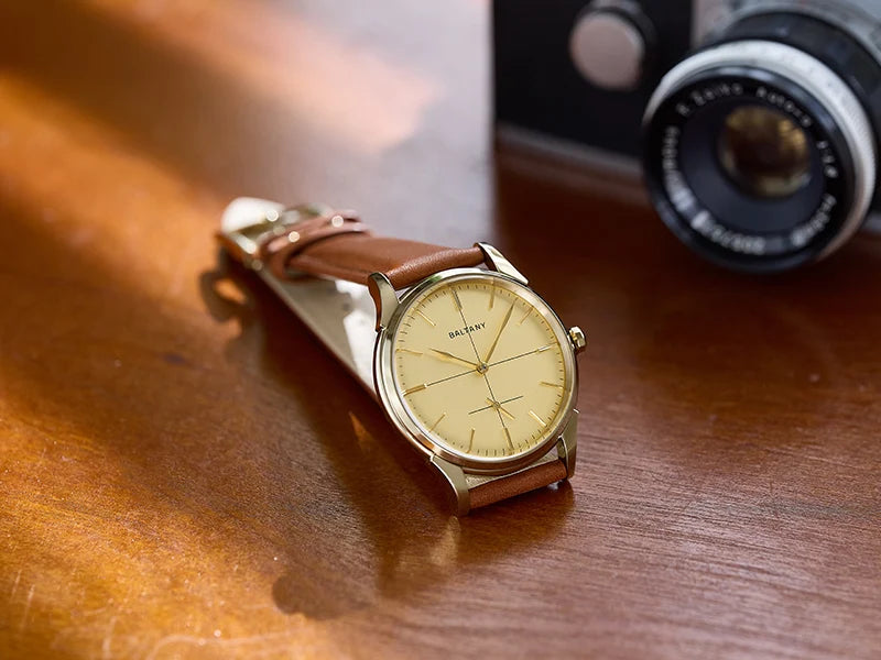 NorthChrono wristwatch featuring a gold-tone case and a tan leather strap, placed on a wooden surface with a vintage camera in the background. The scene is warmly lit, enhancing the classic aesthetic.