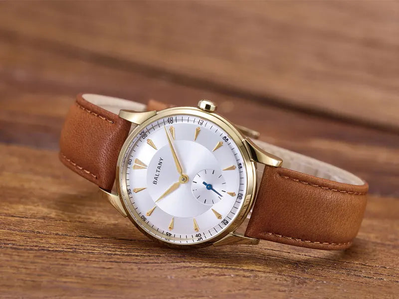 NorthChrono round watch with a white dial featuring gold-toned hour markers and hands, small seconds sub-dial, and a brown leather strap, displayed against a warm wooden background.