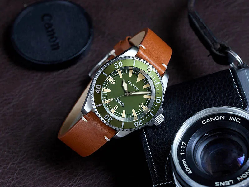 NorthChrono watch featuring a green dial with applied indices, set on a brown leather strap, positioned on a textured surface next to a black Canon camera and lens cap, creating a stylish, artistic setup.
