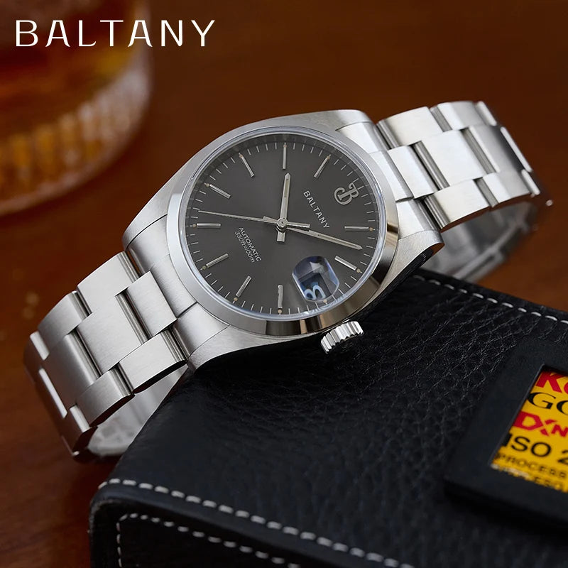 NorthChrono stainless steel wristwatch with a dark gray dial, date function at 6 o'clock, and polished case, displayed on textured black material near blurred background elements suggesting warm beverage. Brand name "BALTANY" visible.