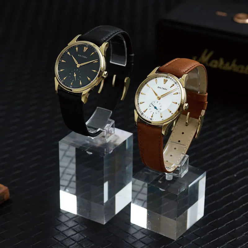 NorthChrono features two wristwatches on clear stands. The left watch has a black dial, gold case, and black leather strap, while the right has a white dial, gold case, and brown leather strap, set against a textured background with a partially visible black box.