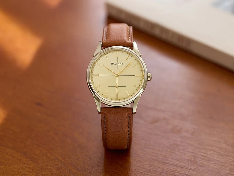 NorthChrono minimalist wristwatch featuring a cream-colored dial with silver hour markers and a brown leather strap, displayed on a wooden surface with a book partially visible in the background.