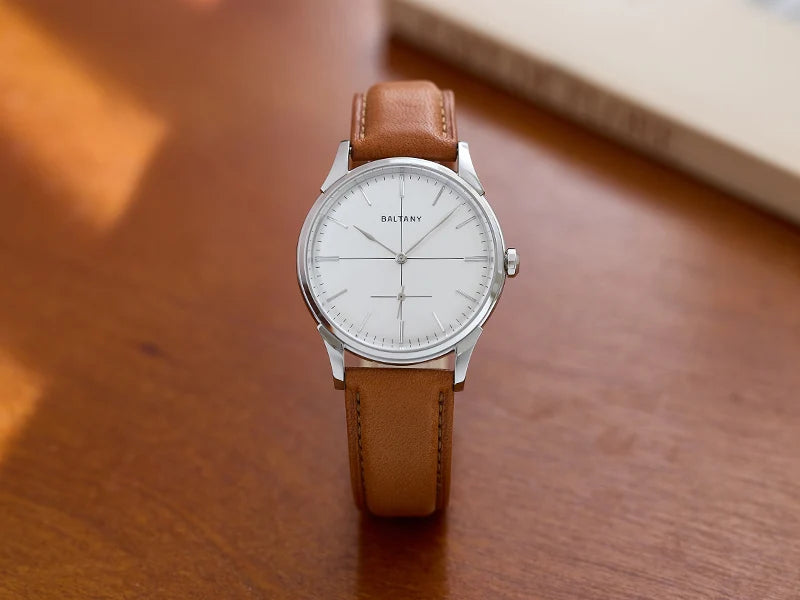 NorthChrono silver minimalist watch on wooden surface, featuring a white dial with thin hour and minute hands, small subdial, and a tan leather strap. A book is partially visible in the background.