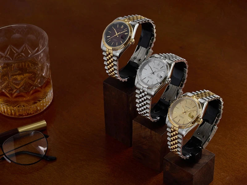 NorthChrono three elegant wristwatches on wooden stands featuring classic designs. The watches have metal bands and different dial colors: one dark, one silver, and one gold. A glass tumbler, a pair of glasses, and a pen are visible in a sophisticated setting.