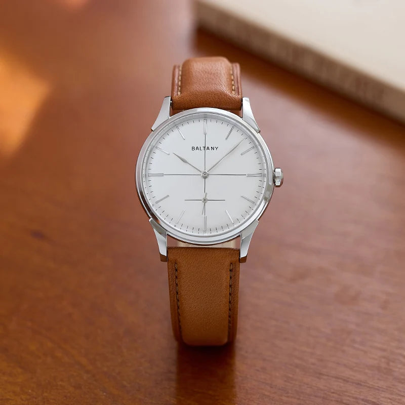 NorthChrono stylish wristwatch on a wooden surface features a white dial with silver hour markers and hands, housed in a sleek silver case. It has a brown leather strap, with a light-colored book visible in the background.