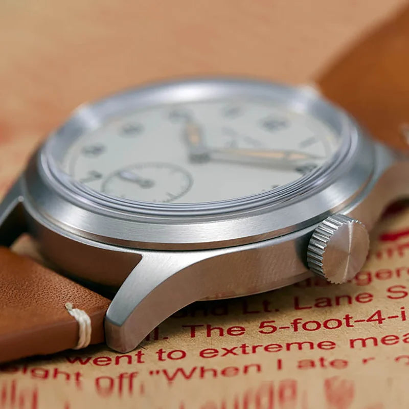 NorthChrono close-up of a round stainless steel watch featuring a white dial with silver applied indices, luminous hands, and a subdial. The watch is on a tan leather strap, set against a textured background with visible printed text.