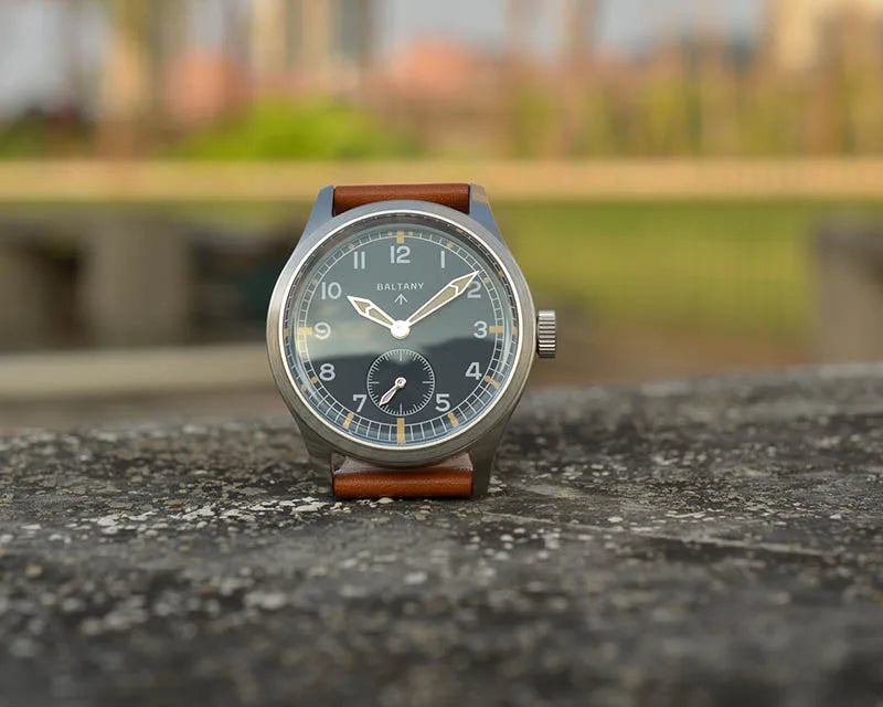 NorthChrono round metal case wristwatch with a dark dial featuring white hour markers and hands, a small sub-dial near the bottom, and a brown leather strap, set against a blurred outdoor background with greenery.