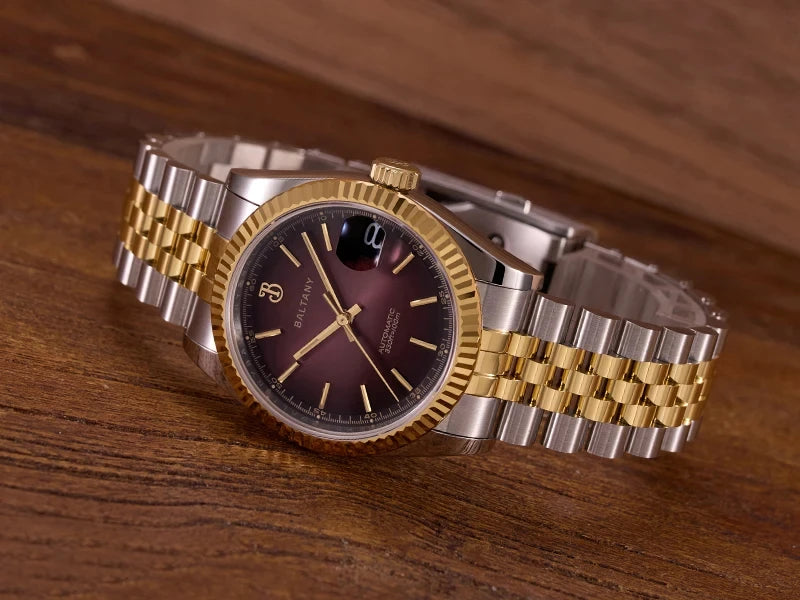 NorthChrono elegant two-tone wristwatch featuring a round face with a gradient maroon dial, gold hour markers, and hands. It has a date display at three o'clock and a fluted bezel, set on a wooden surface.