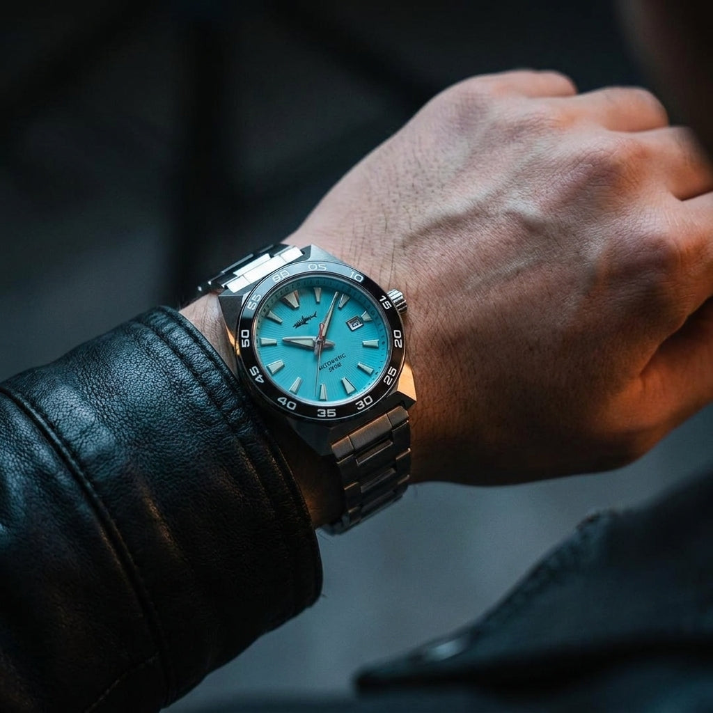 NorthChrono close-up of a watch featuring a bright turquoise dial with applied indices and luminous hands, paired with a metallic strap. The hand is adorned with a black leather jacket sleeve against a softly blurred background.