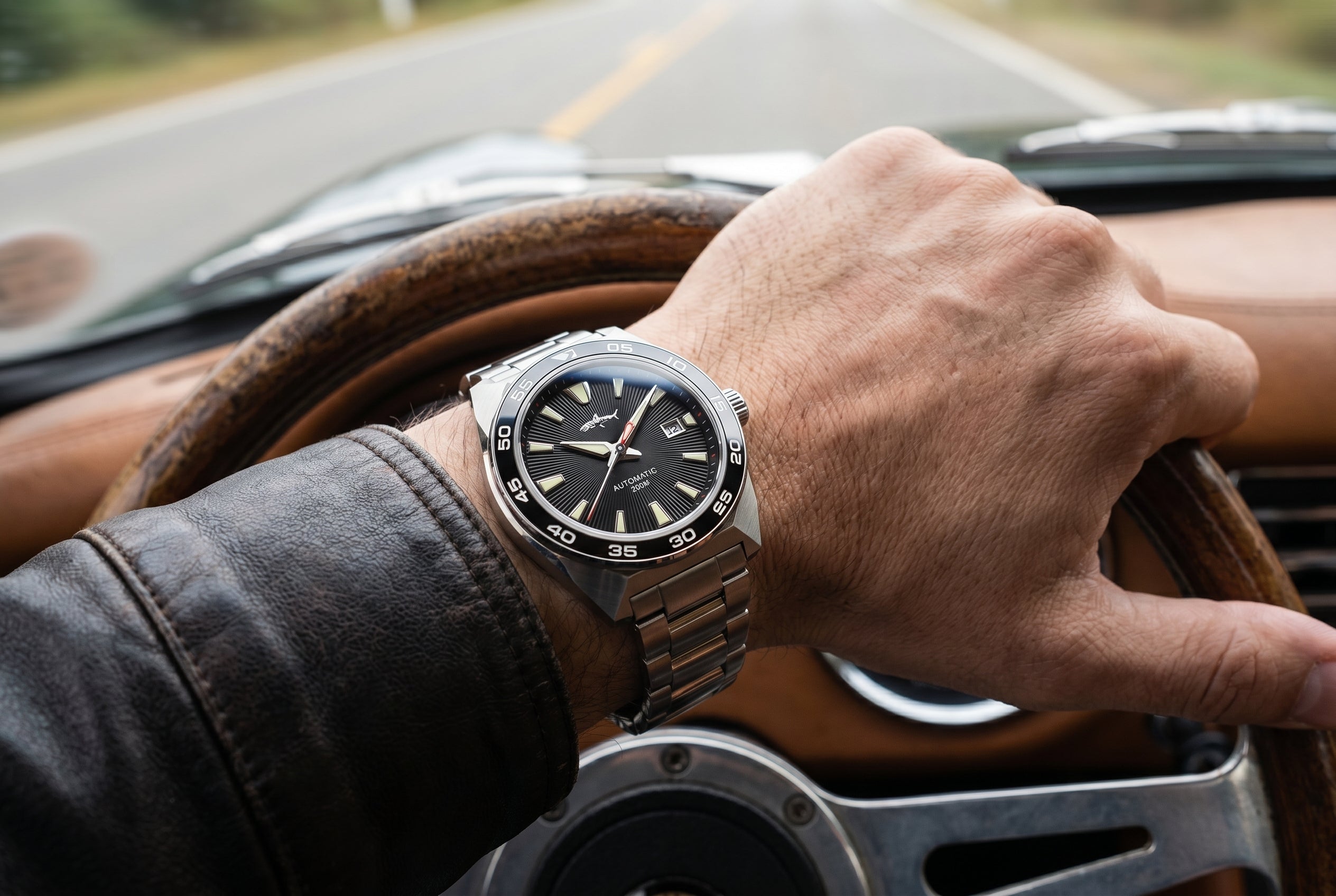 NorthChrono close-up of a male wrist wearing a stainless steel watch with a black dial and silver accents, gripping a steering wheel inside a car, with a scenic road view in the background.