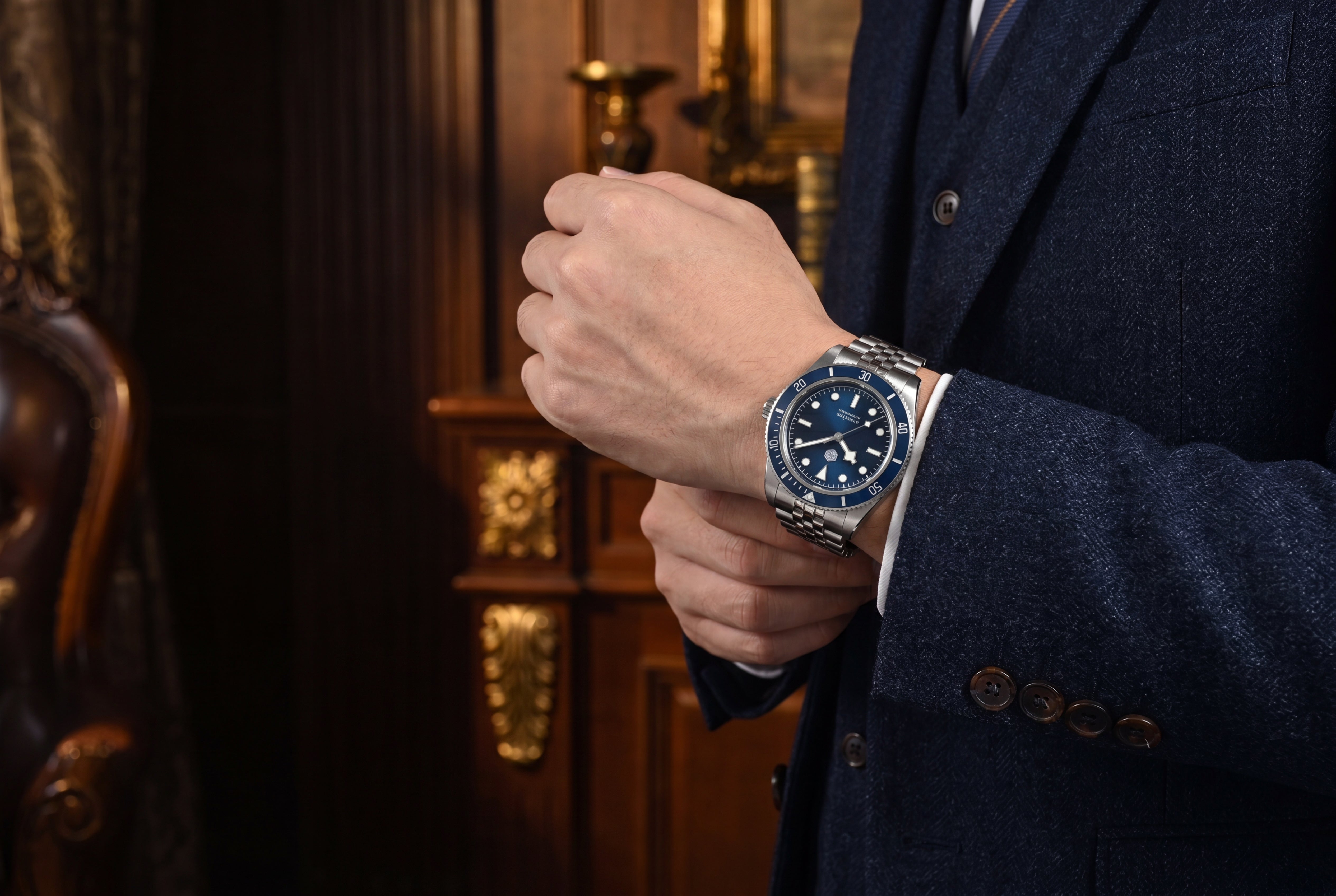 NorthChrono close-up of a wrist wearing a watch with a blue dial, applied indices, and luminous hands, complemented by a metallic bracelet. The person is dressed in a dark suit, set against an elegant background.